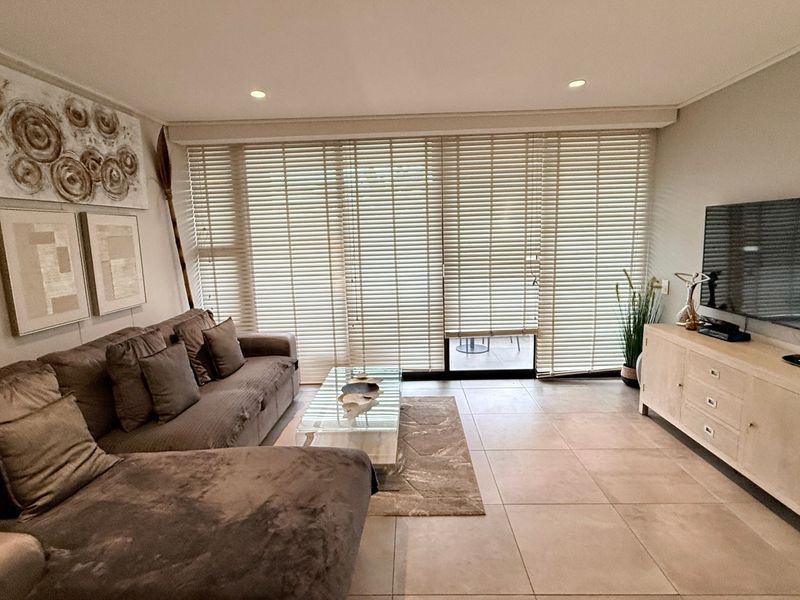 A well-maintained living room featuring neutral tiled flooring, recessed downlighting, and large sliding glass doors fitted with venetian blinds for natural light control.