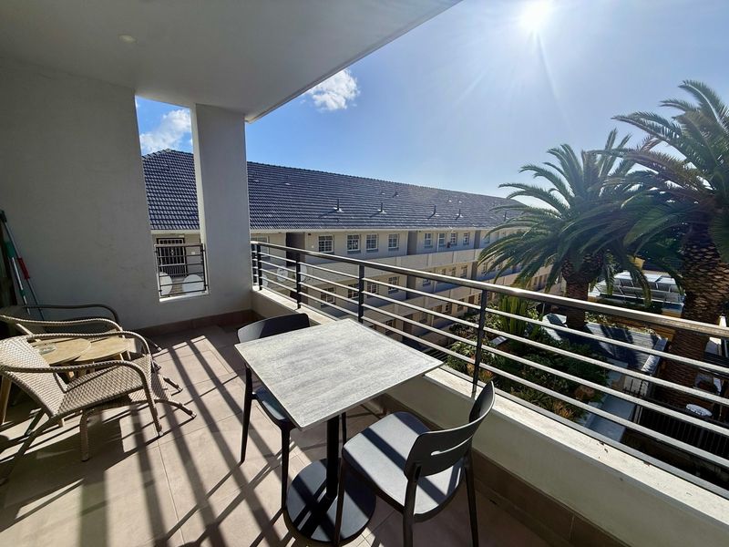 A well-maintained covered balcony featuring tiled flooring and a metal balustrade, offering an open view of the surrounding apartment complex.
