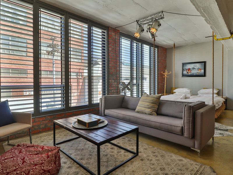 Spacious open-plan apartment featuring industrial-style exposed brick walls, polished concrete floors, and large windows with metal shutters, creating a bright and modern living space.