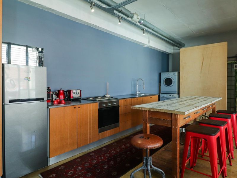 Industrial-style kitchen featuring wooden cabinetry, stainless steel countertops, built-in oven, cooktop, and integrated laundry area with exposed ceiling services.