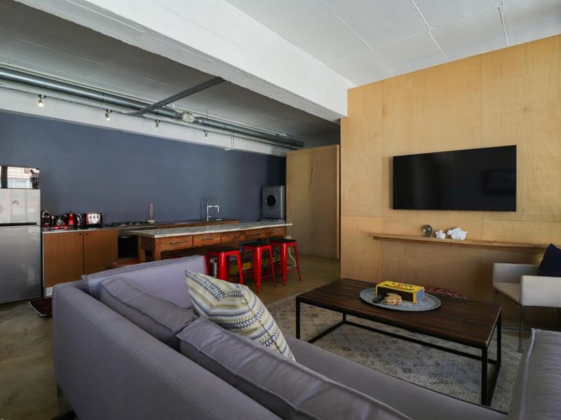 Modern open-plan living area featuring industrial-style exposed ceilings, plywood wall paneling, concrete floors, and a functional kitchen with built-in cabinetry.