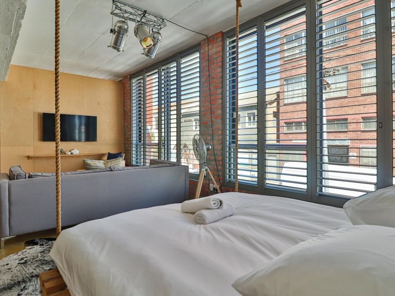 A modern industrial-style bedroom featuring exposed brick walls, plywood paneling, concrete ceilings, and large windows with integrated shutters.