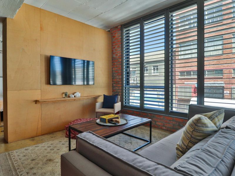 Modern industrial-style living room featuring exposed brick walls, plywood paneling, polished concrete floors, and large windows providing ample natural light.