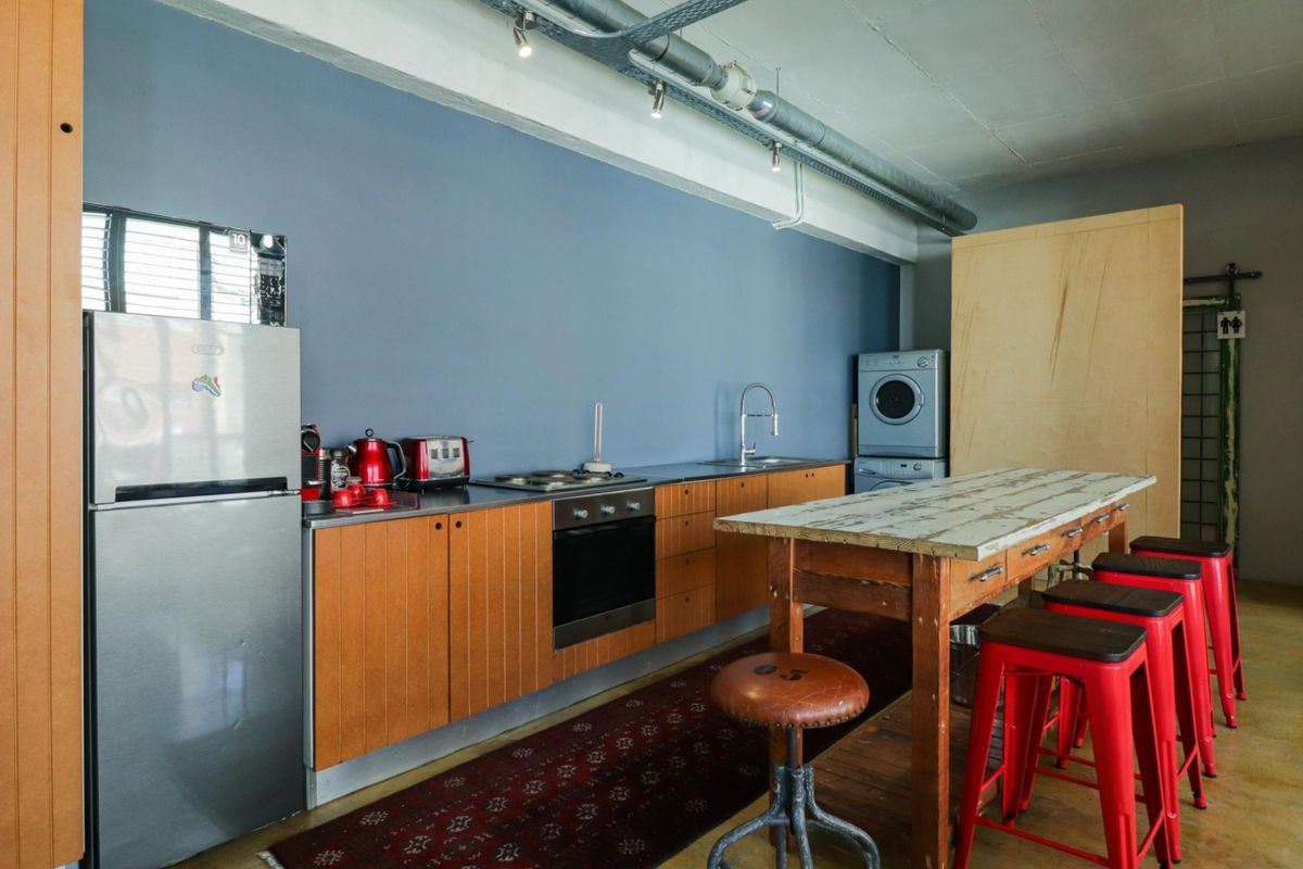 Industrial-style kitchen featuring wooden cabinetry, stainless steel countertops, built-in oven, cooktop, and integrated laundry area with exposed ceiling services.