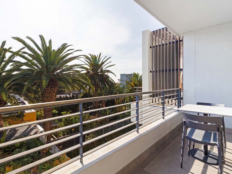 A well-maintained balcony featuring a metal balustrade, tiled flooring, and a vertical privacy screen, offering a pleasant outlook over palm trees.