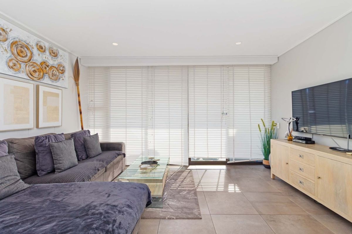 A bright and modern living room featuring large format floor tiles, recessed downlighting, and floor-to-ceiling venetian blinds leading to an outdoor area.