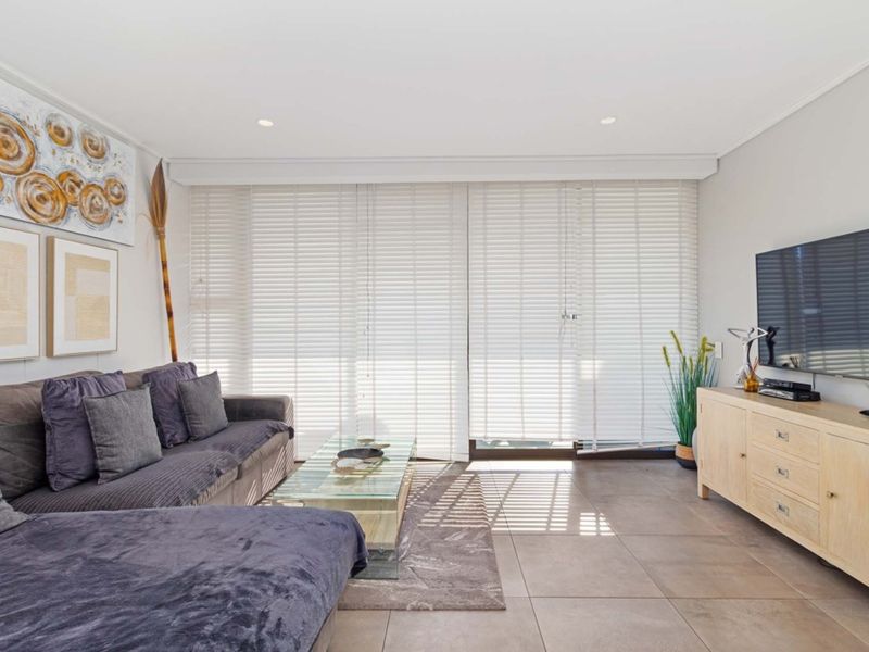 A bright and modern living room featuring large format floor tiles, recessed downlighting, and floor-to-ceiling venetian blinds leading to an outdoor area.