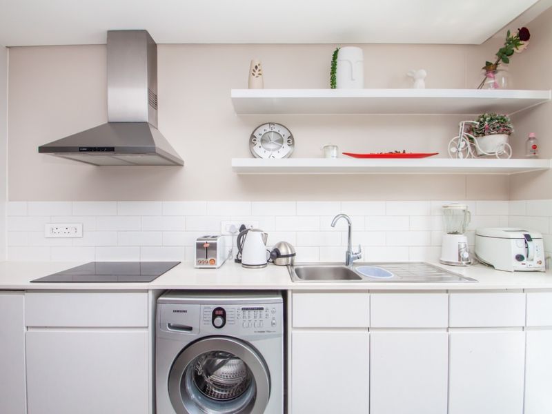 Modern kitchen featuring white cabinetry, subway tile backsplash, stainless steel extractor fan, electric hob, and an integrated washing machine with floating shelving.