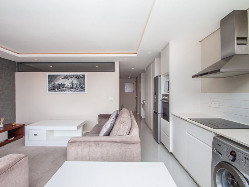 Modern open-plan living area featuring a sleek kitchen with integrated appliances, subway tile backsplash, and recessed ceiling lighting, flowing into a comfortable lounge space.