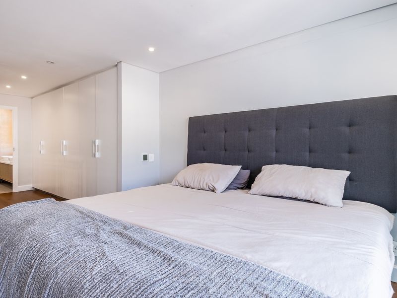 A well-maintained bedroom featuring built-in cupboards, recessed downlighting, and hardwood flooring, with an adjoining en-suite bathroom visible in the background.