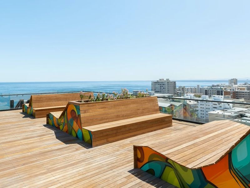 A modern rooftop terrace featuring timber decking, built-in seating with artistic detailing, and glass balustrades offering expansive ocean and city views.