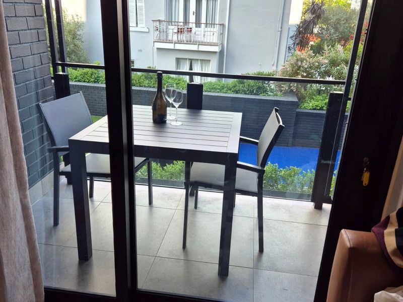 A modern balcony featuring a glass balustrade, tiled flooring, and a dark brick feature wall, overlooking a communal pool area.