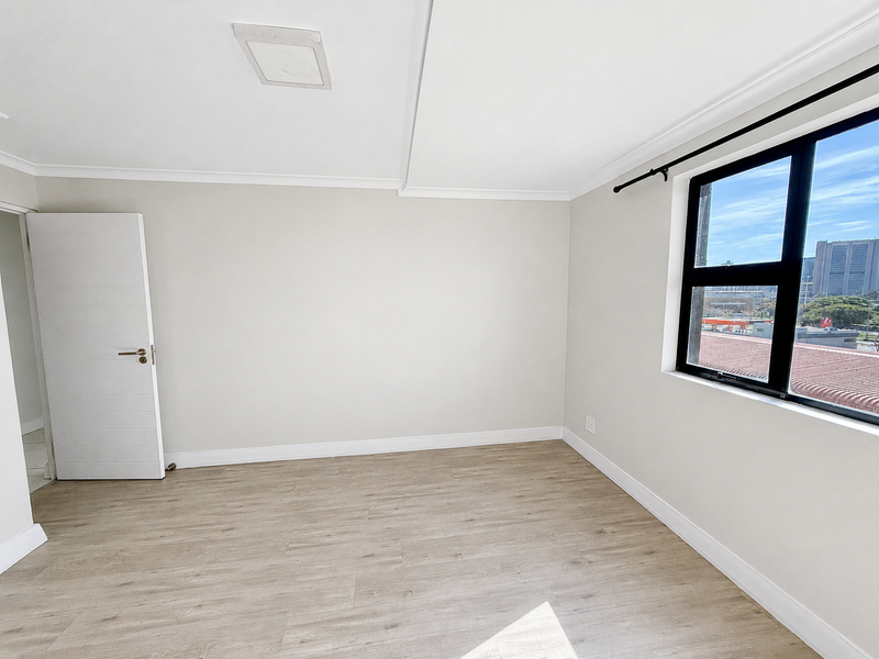 A bright, empty bedroom featuring modern laminate flooring, neutral walls, and a large window with views of the surrounding area.
