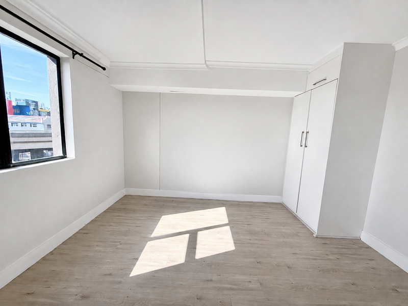 A bright, empty bedroom featuring light-toned laminate flooring, neutral walls, and a built-in wardrobe with modern hardware.