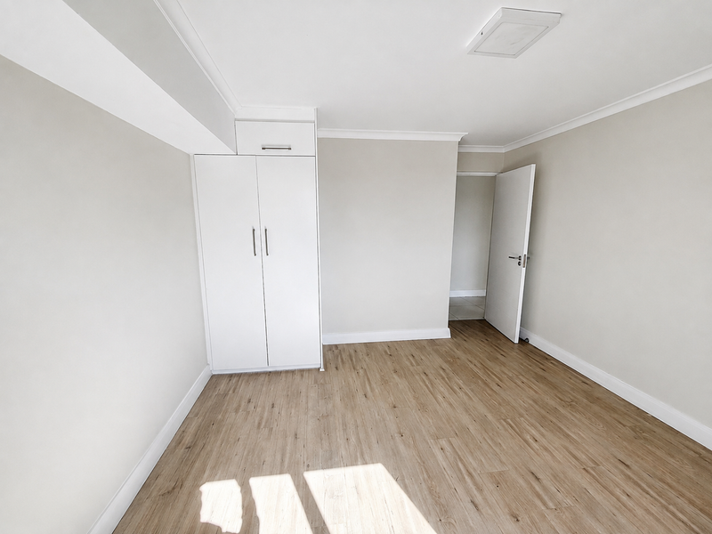 A bright, empty bedroom featuring neutral walls, modern laminate flooring, and a built-in wardrobe with overhead storage.