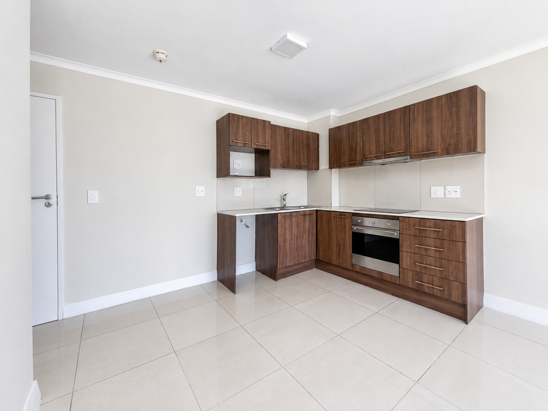 A clean, modern kitchen featuring dark wood cabinetry, integrated appliances, and neutral tiled flooring in an open-plan layout.