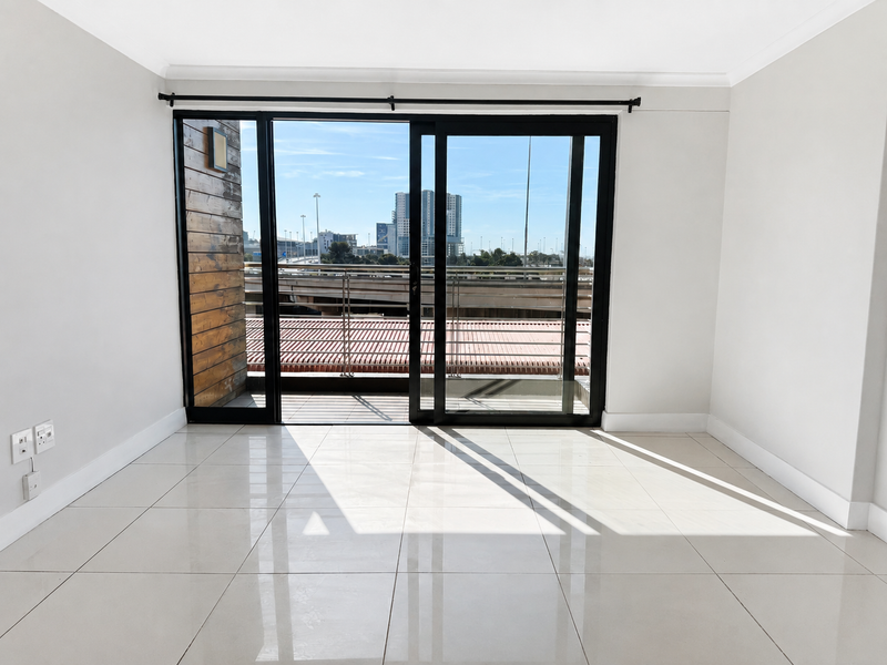 A bright, empty living room featuring large sliding glass doors that open onto a balcony, with polished floor tiles and neutral wall finishes.