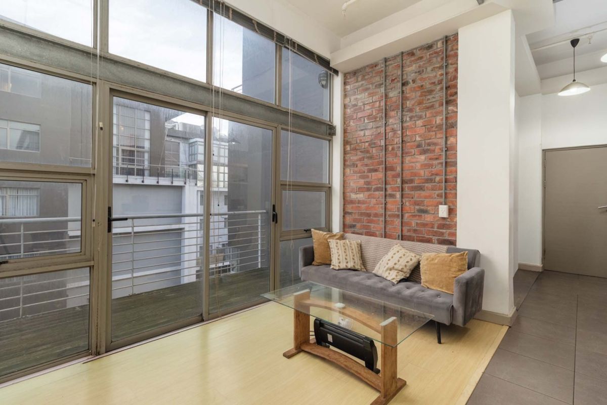 A bright living area featuring an industrial-style exposed brick wall, high ceilings, and large floor-to-ceiling windows providing access to a private balcony.