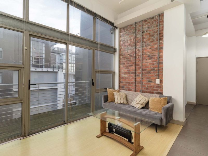 A bright living area featuring an industrial-style exposed brick wall, high ceilings, and large floor-to-ceiling windows providing access to a private balcony.
