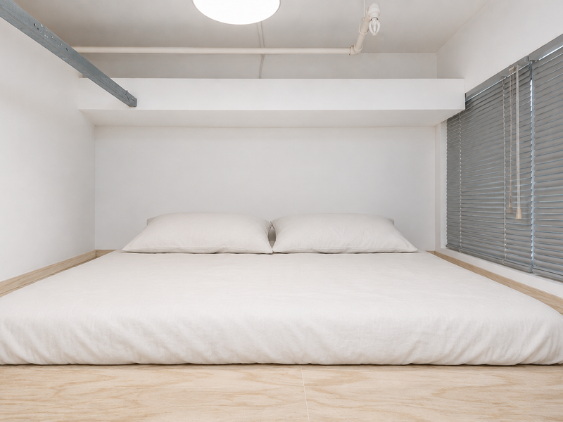 A minimalist bedroom featuring light wood flooring, venetian blinds, and an exposed ceiling pipe, creating a clean and modern aesthetic.