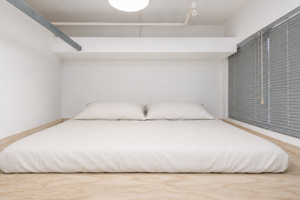 A minimalist bedroom featuring light wood flooring, venetian blinds, and an exposed ceiling pipe, creating a clean and modern aesthetic.