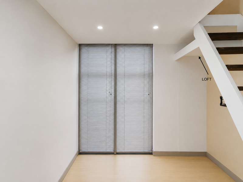 A bright, empty living area featuring laminate flooring, recessed lighting, venetian blinds, and a staircase leading to a loft space.