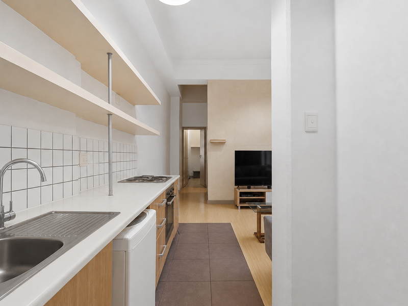 A functional galley-style kitchen featuring light wood cabinetry, open shelving, a tiled backsplash, and integrated cooking appliances, flowing into the adjacent living area.