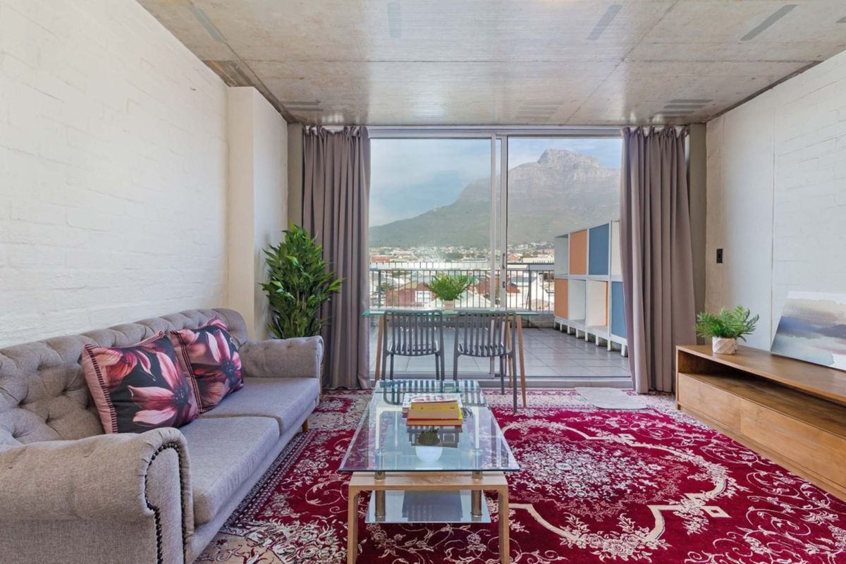 A bright living room featuring an exposed concrete ceiling, painted brick walls, and large sliding doors opening onto a balcony with mountain views.