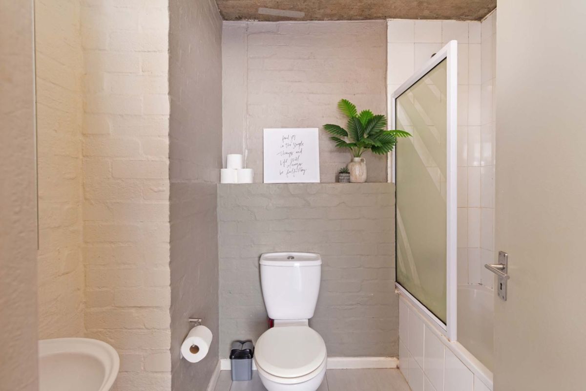 A functional bathroom featuring a toilet, bathtub with a shower screen, and a combination of tiled and painted brick wall finishes.