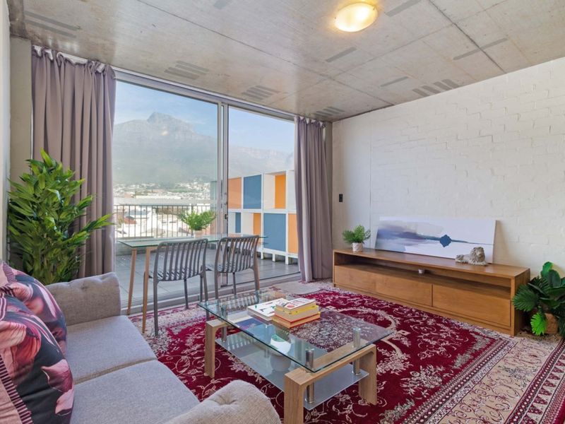 A bright living room featuring an exposed concrete ceiling, painted brick wall, and large sliding glass doors opening onto a balcony with mountain views.