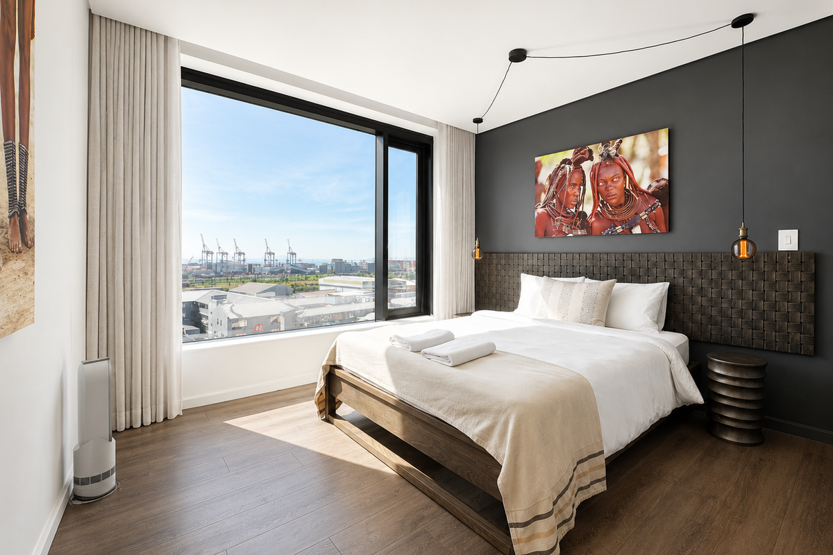 A modern, well-lit bedroom featuring large floor-to-ceiling windows with expansive city and industrial views, high-quality laminate flooring, and a contemporary dark accent wall.