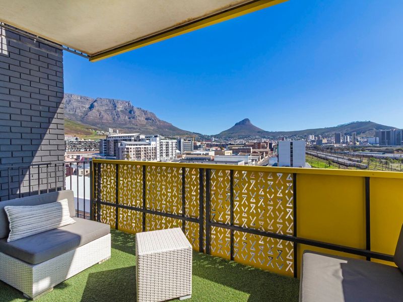 A modern balcony featuring a decorative yellow metal balustrade, artificial grass flooring, and expansive views of the city and mountains.
