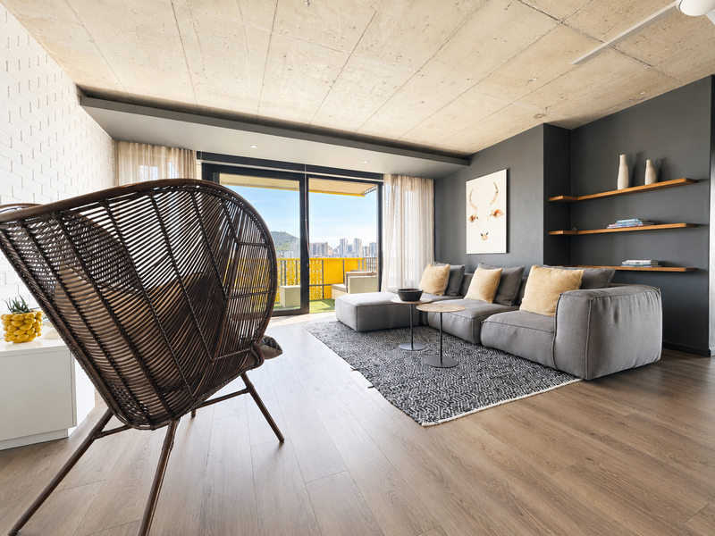 Modern living room featuring an exposed concrete ceiling, timber laminate flooring, built-in shelving, and large sliding doors opening to a balcony with city views.