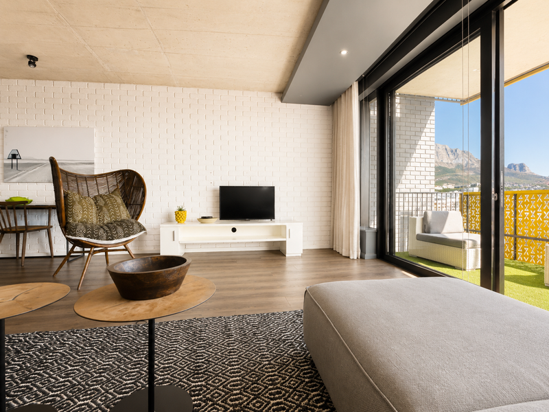 Modern living room featuring an exposed concrete ceiling, painted brick feature wall, and large sliding glass doors opening onto a balcony with mountain views.
