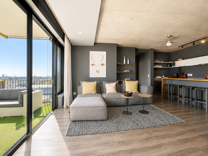 Modern open-plan living area featuring exposed concrete ceilings, laminate flooring, and a sleek kitchen with a breakfast bar, flowing onto a private balcony.