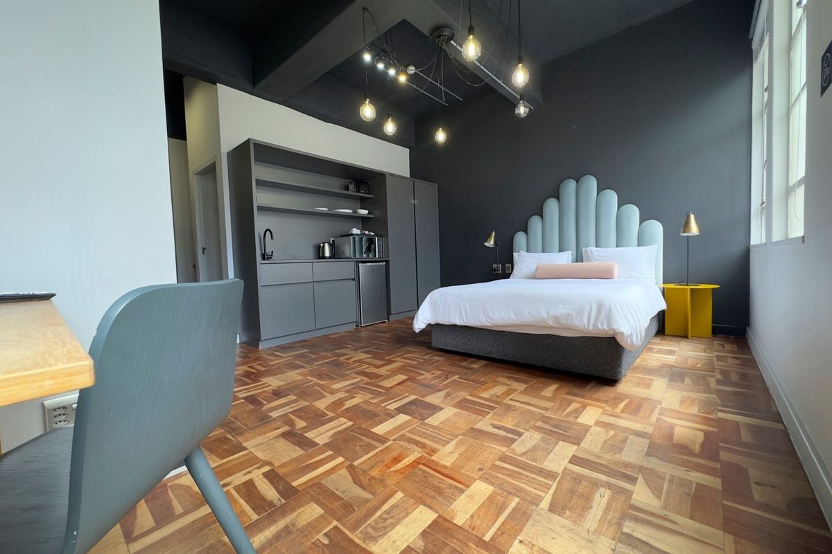 A modern bedroom featuring parquet flooring, exposed ceiling beams, and a convenient built-in kitchenette area with dark cabinetry.