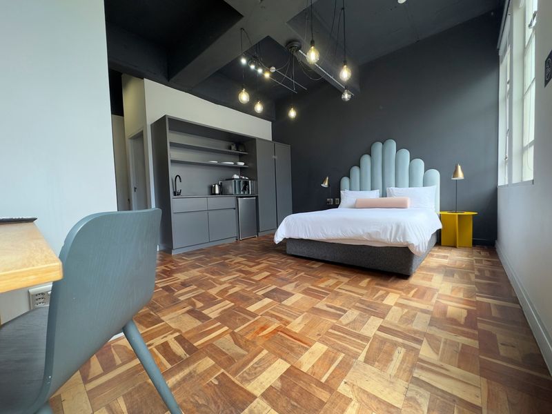 A modern bedroom featuring parquet flooring, exposed ceiling beams, and a convenient built-in kitchenette area with dark cabinetry.