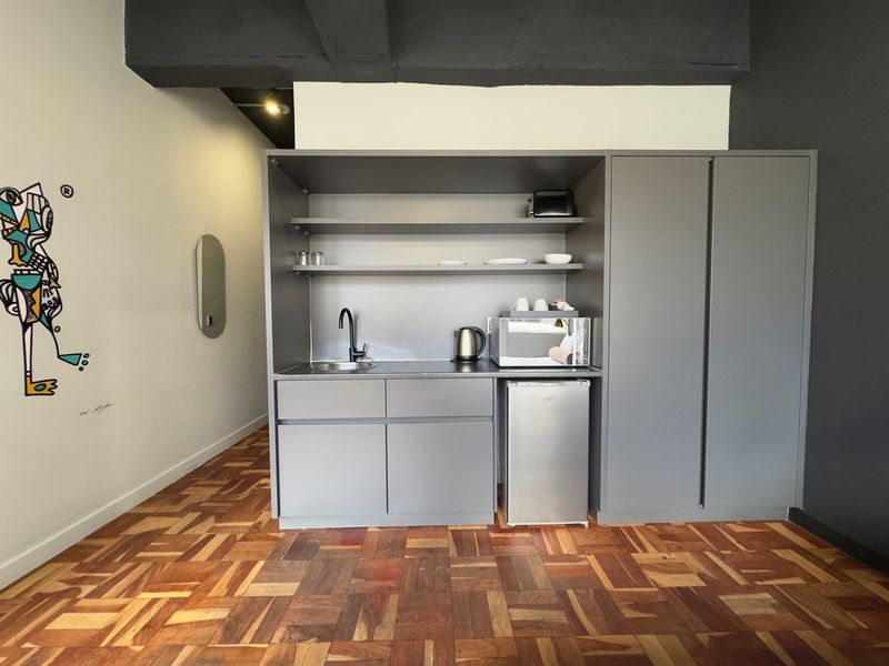 A compact kitchenette featuring grey built-in cabinetry, open shelving, a sink, and warm-toned parquet flooring in a modern apartment setting.