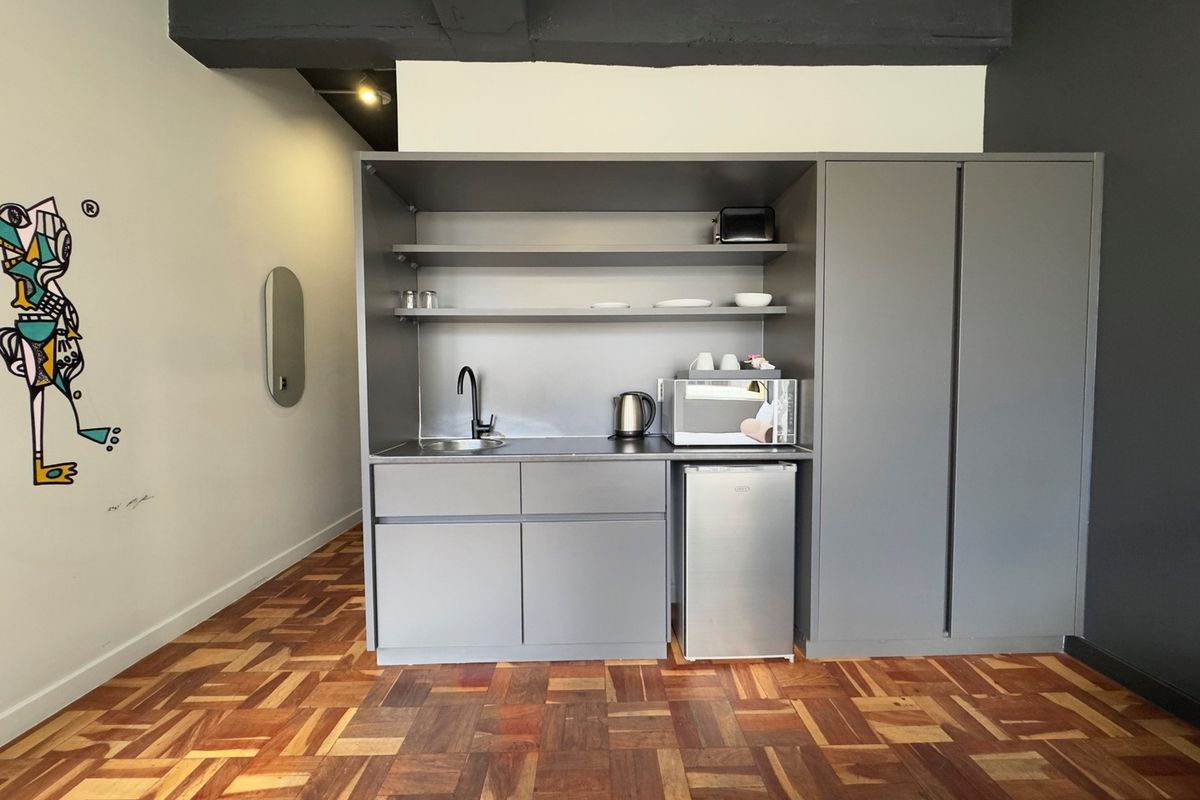 A compact kitchenette featuring grey built-in cabinetry, open shelving, a sink, and warm-toned parquet flooring in a modern apartment setting.