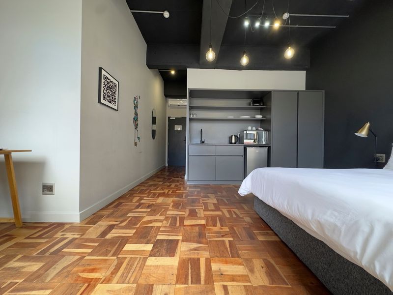 Modern open-plan studio apartment featuring parquet flooring, a compact built-in kitchenette, and stylish exposed ceiling beams with pendant lighting.
