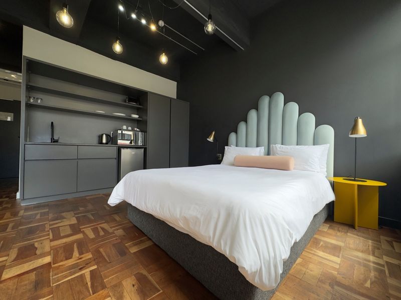Modern bedroom featuring dark walls, parquet flooring, exposed ceiling beams, and a compact built-in kitchenette area with integrated storage.