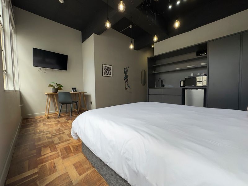 Modern bedroom featuring parquet flooring, exposed dark ceiling beams, and a compact built-in kitchenette area with integrated cabinetry and sink.