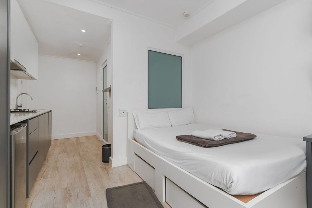 A compact open-plan studio apartment featuring a modern kitchenette with built-in cabinetry and a space-saving bed frame with integrated storage.