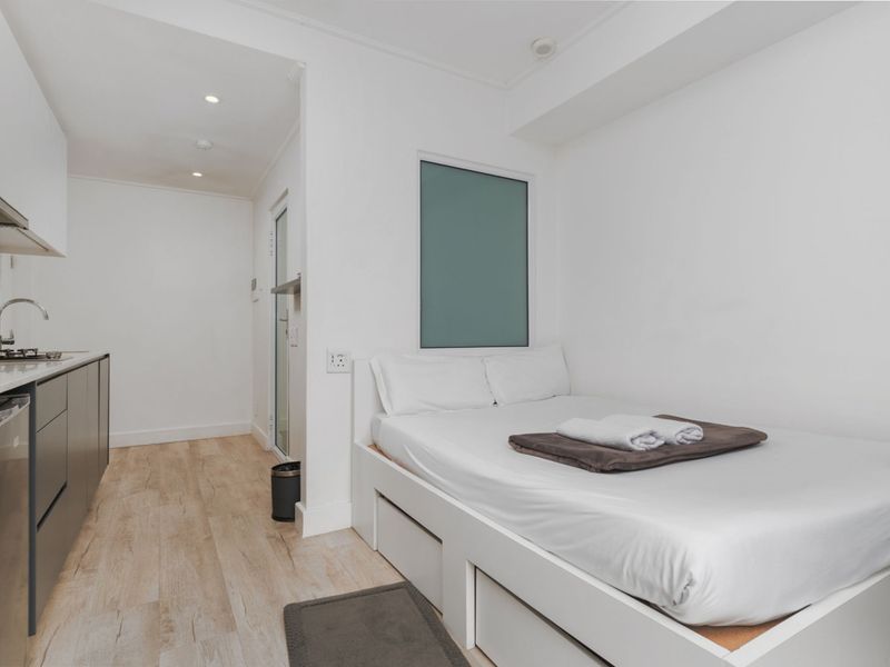 A compact open-plan studio apartment featuring a modern kitchenette with built-in cabinetry and a space-saving bed frame with integrated storage.