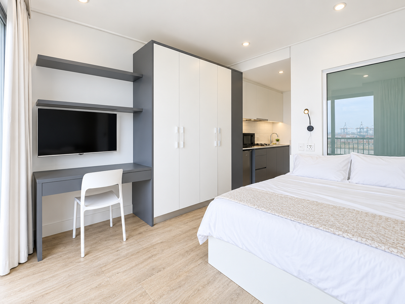 Modern open-plan studio apartment featuring a sleek kitchen, built-in wardrobe, integrated desk area, and light wood-look flooring with contemporary finishes.