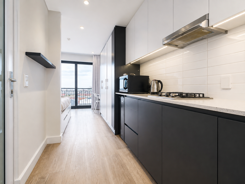 Modern open-plan apartment kitchen featuring sleek dark cabinetry, stone countertops, subway tile backsplash, and integrated gas hob, leading to a balcony with city views.