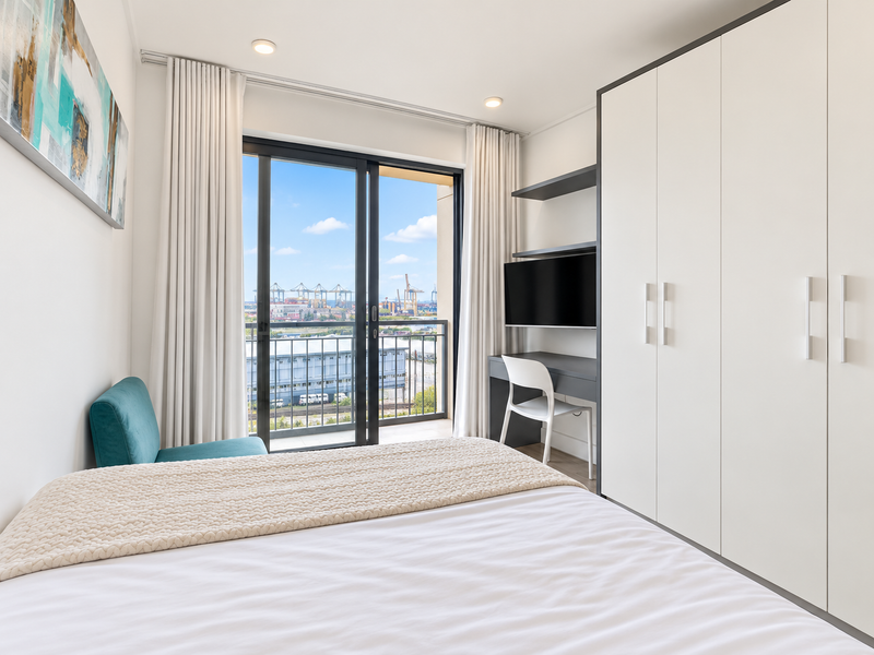 Modern bedroom featuring built-in wardrobes, a integrated desk workspace, and sliding glass doors leading to a balcony with an industrial view.