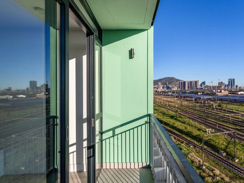 A modern balcony featuring a metal railing and sliding glass door access, offering expansive views of the city skyline and railway infrastructure.