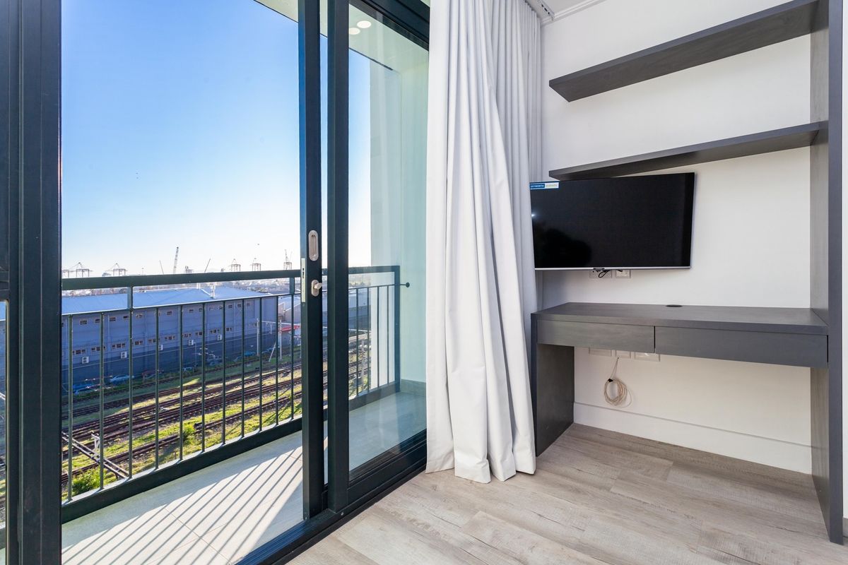 Modern bedroom featuring built-in desk and shelving, with a sliding glass door leading to a private balcony offering an industrial view.
