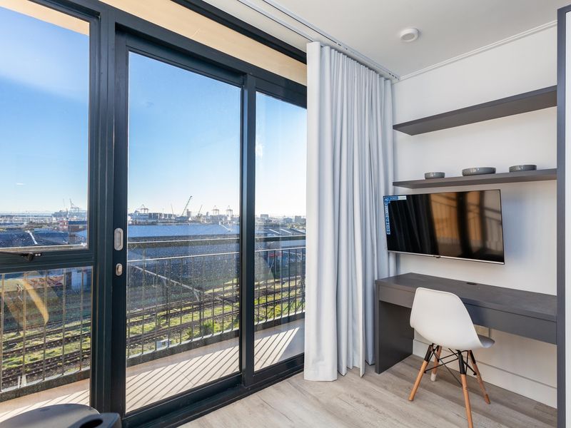 Modern bedroom featuring built-in desk and shelving, laminate flooring, and large sliding glass doors opening to a balcony with industrial views.